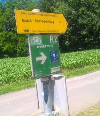 A yellow hiking finger sign on a post above a square green cycling sign with a white arrow