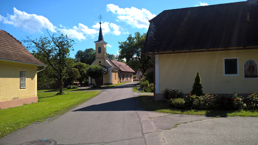 Small road passing between a house and a small neat chapel with a little bell tower and pointy black spire.