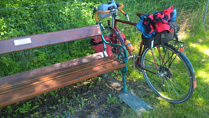 Front end of bike with red front panniers leaning up against the green cast-iron end of a roadside bench.