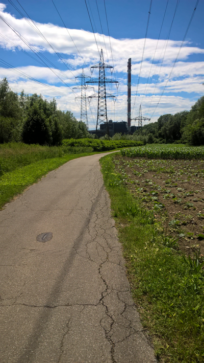 Path between fields, power lines overhead, high pylons, the dark shape of a power station and a tall chimney ahead.