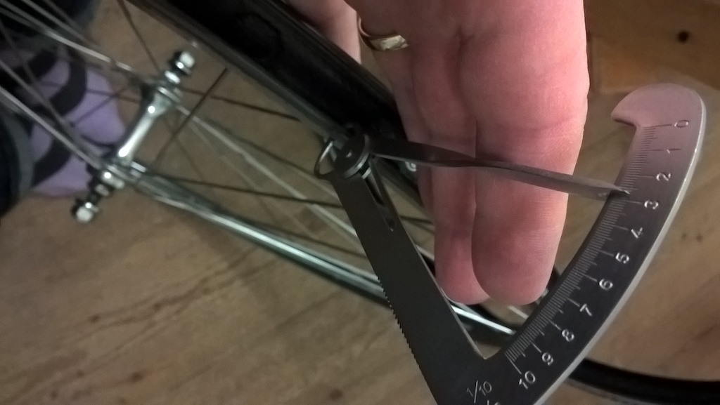 Edge of a bicycle wheel with the tyre off; a hand is holding the Iwanson gauge so that it grips the rim below the bead.