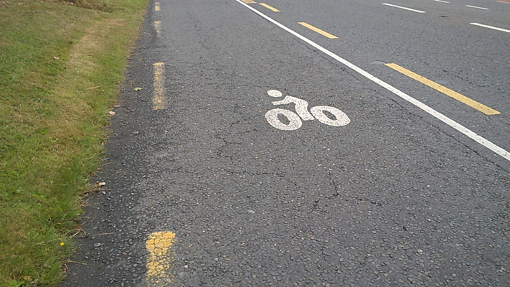 Edge of a main road with a small bicycle pictogram marked on the road. Both yellow hard shoulder and white bike lane markings are present.
