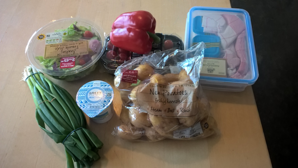 A collection of food on a table: spring onions, some salad, a pepper, a platic box with things inside, a transparent bag of small potatoes and a small carton of yoghurt