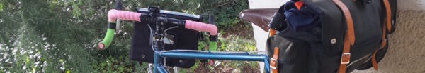 top of bike. handlebar tape is a mixture of pink and green, saddlebag is full, to the left, a dark green conifer in the background, bike is leaning against a low cream coloured wall