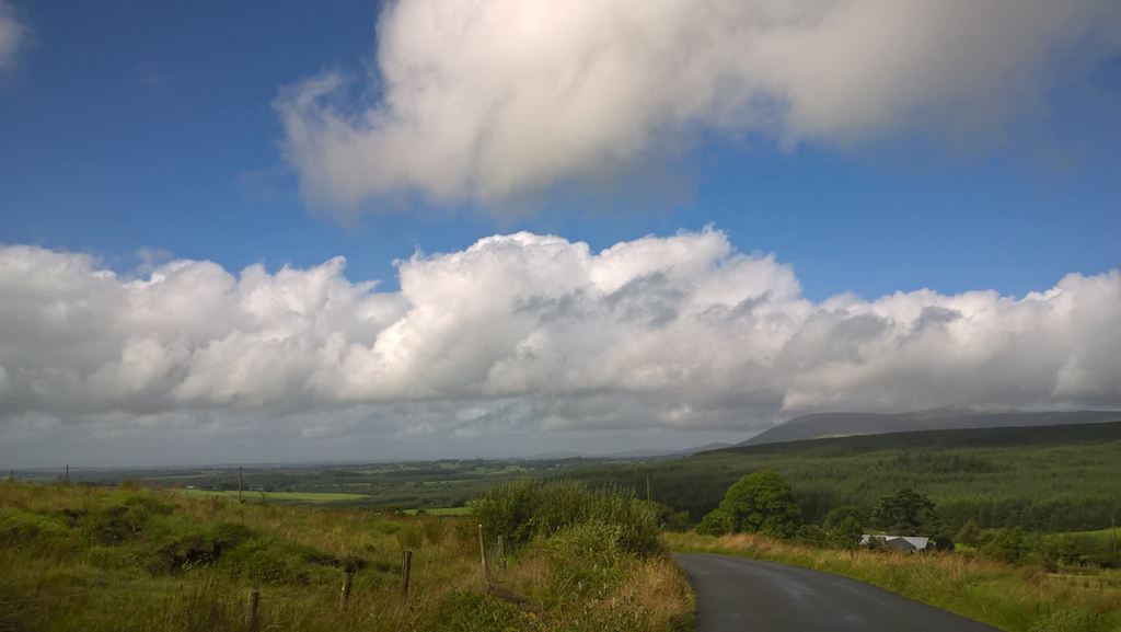 Bright blue sky over a wide green landscape: rows of fluffy white clouds in the background