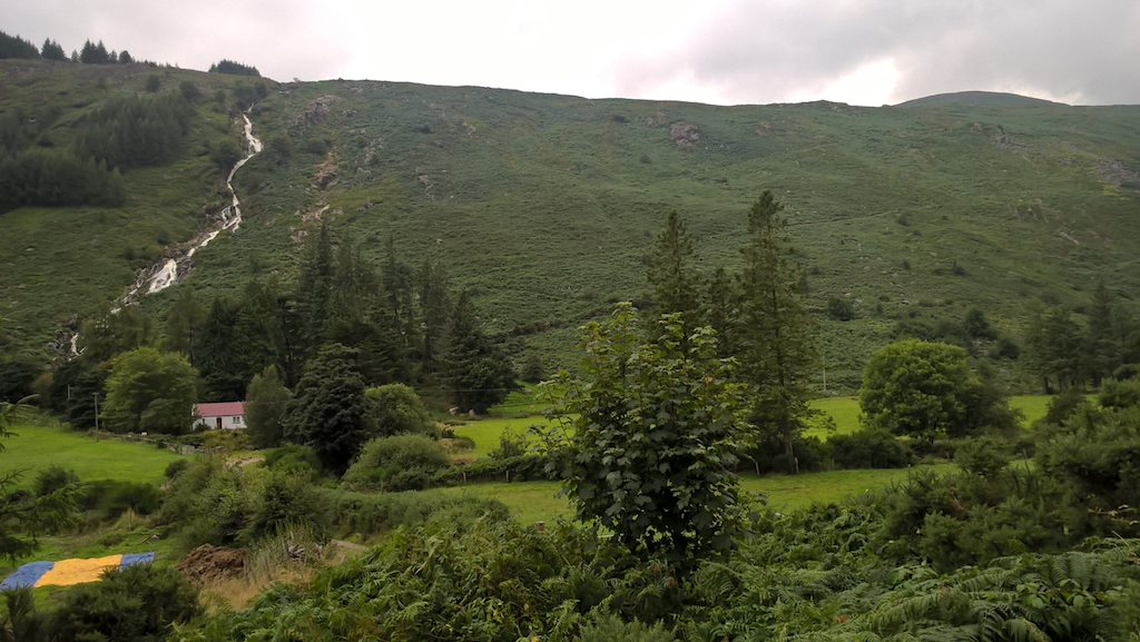 green hillside with fields in foreground; small groups of trees; behind the fields, a house with a red roof, behind the house, steeply rising moorland with a waterfall
