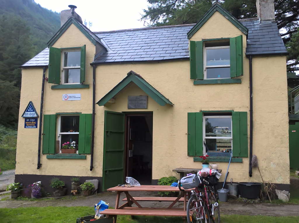 A two-storey house, one window either side of the door and one dormer window above each ground-floor window, windows and doors are green, facade painted cream, a slate roof, bike leaning against picnic table in front