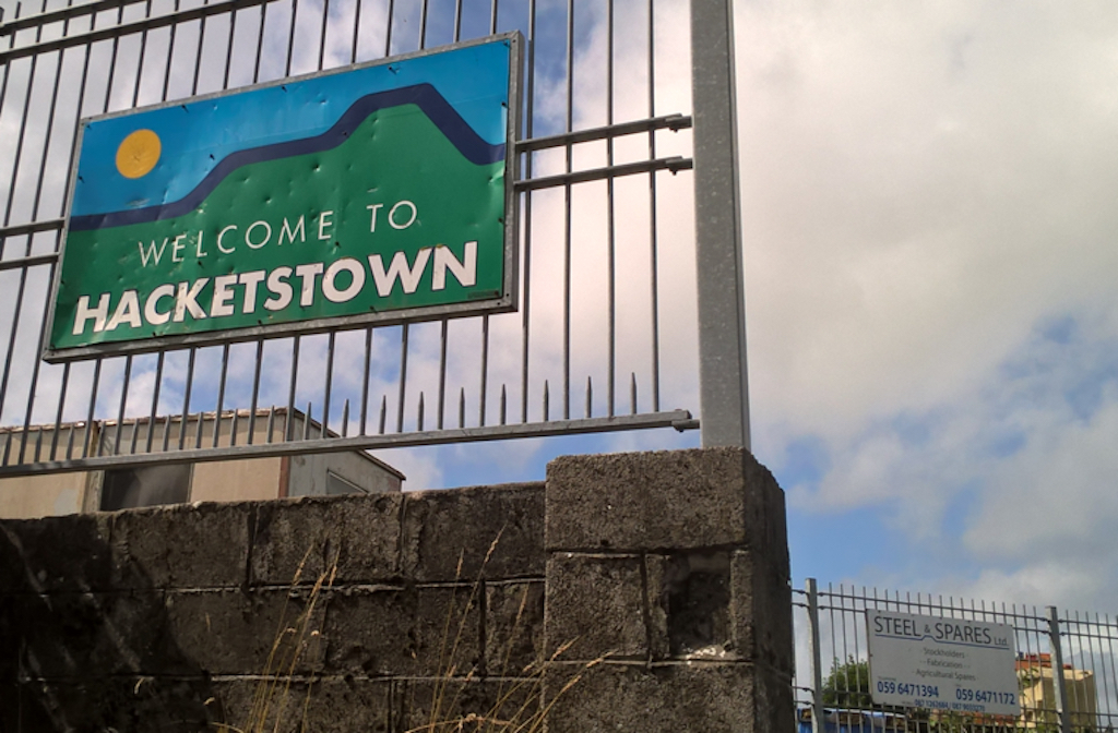 Sign "Welcome to Hacketstown" on a metal fence with blue sky and white clouds behind