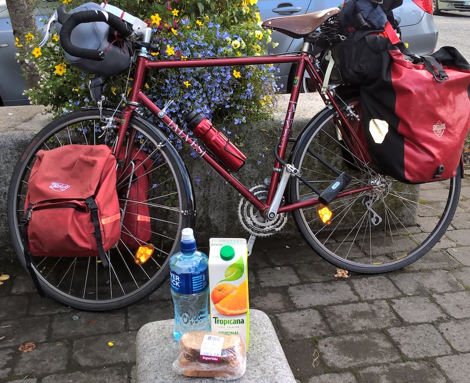 Loaded touring bike parked against a stone bench with flowers; water, ourange juice and sandwiches in front