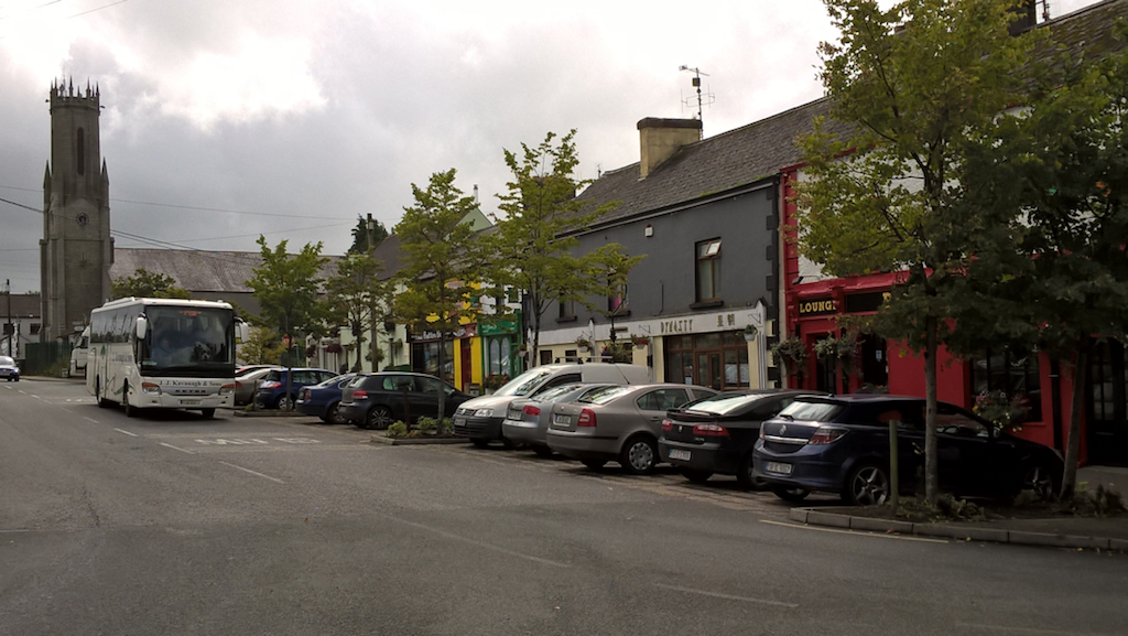 Wide village street in Ireland with pubs and shops, a church in the background and a bus stopping