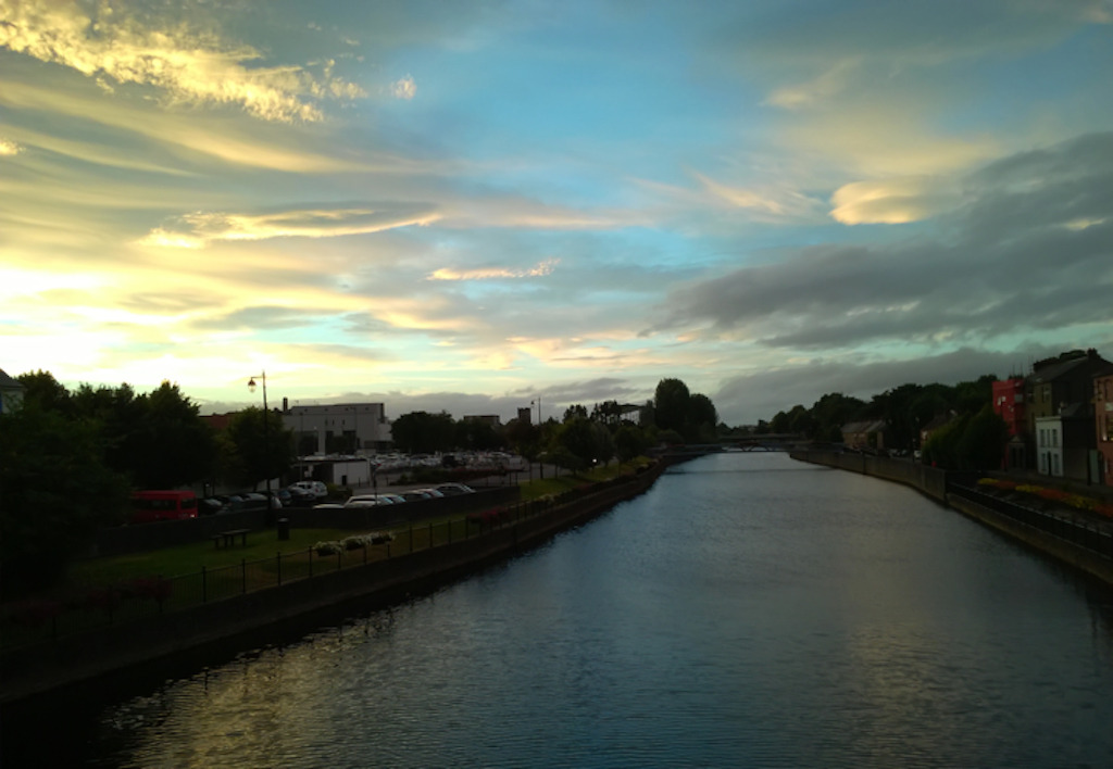 Sun setting with golden clouds and blue sky over a river, low buildings on both sides