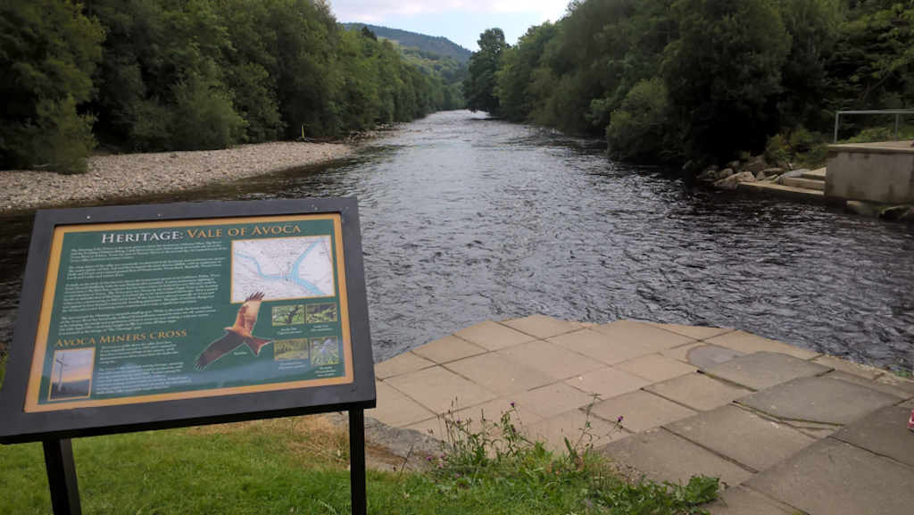 A paved point of river bank at the confluence of two rivers, looking downstream; banks are wooded; a sign explains the situation