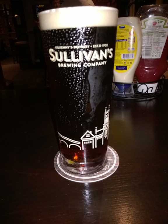 A pint of Sullivan's stout, dark brown with a white head, condensation and a slight overflow on the glass
